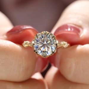 May include: A gold engagement ring with a large, round, clear gemstone. The ring has an ornate, floral-inspired setting with intricate detailing. The ring is held between two hands.