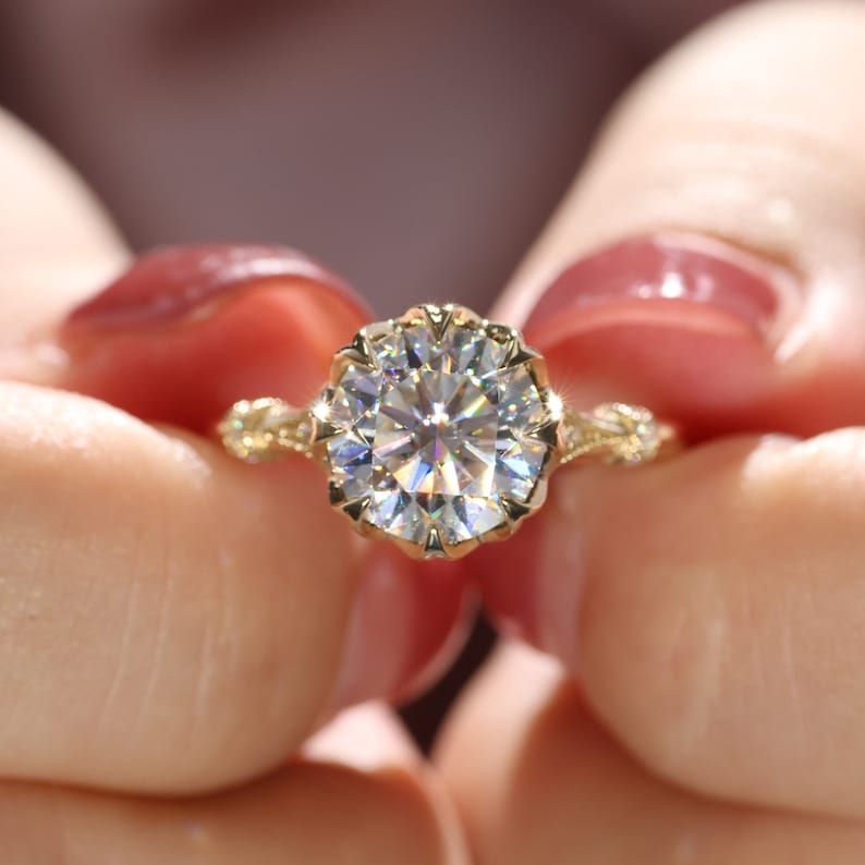 May include: A gold engagement ring with a large, round, clear gemstone. The ring has an ornate, floral-inspired setting with intricate detailing. The ring is held between two hands.