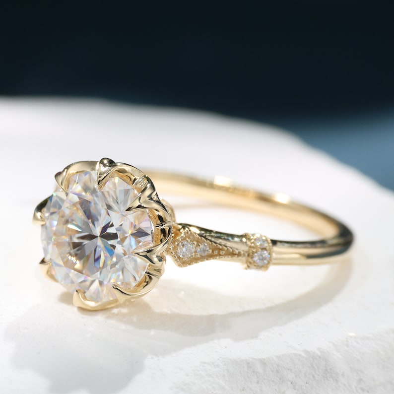 May include: A gold engagement ring with a large, round, clear gemstone. The ring features a floral-inspired setting and small accent stones on the band. The ring is set against a white surface with a dark blue background.