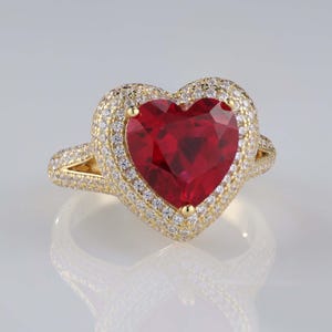 May include: A gold ring featuring a large, heart-shaped, faceted red gemstone. The gemstone is surrounded by two rows of smaller, clear gemstones. The ring's band and setting are gold-colored.