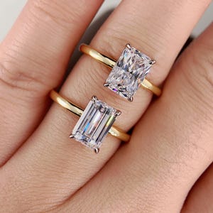 May include: Two gold rings each feature a rectangular, clear gemstone. The rings are set on a gold band. The gemstones are held in place by four prongs. The rings are displayed on a light skin tone.