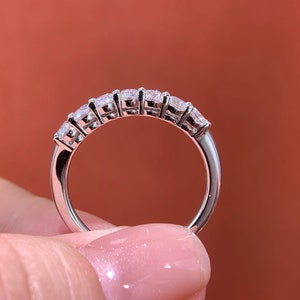 May include: A silver ring with seven clear, square-cut gemstones set in a row. The ring is displayed against a warm, reddish-brown backdrop, highlighting the sparkling stones and the polished metal band.