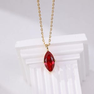 Op de afbeelding: Een goudkleurige ketting met een marquise-geslepen, dieprode edelsteen hanger. De hanger hangt aan een fijne ketting. De ketting wordt gepresenteerd op een wit oppervlak, waardoor een contrast ontstaat.