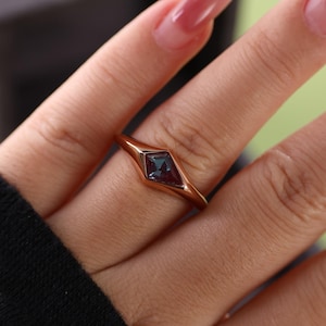 May include: A rose gold ring featuring a square-cut, dark blue-green gemstone. The ring has a sleek, minimalist design with a slightly raised bezel setting. The band is smooth and polished, creating a modern and elegant look.