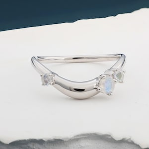 May include: A curved silver ring with three small, round, iridescent gemstones. The ring has a smooth, polished finish and a modern, minimalist design. The gemstones are set along the curve of the ring.