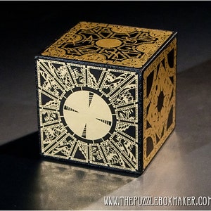 May include: A black and gold puzzle box with intricate geometric designs. The cube features a circular design on one side and detailed patterns across its faces. The text "THEPUZZLEBOXMAKER.COM" is visible on the bottom.