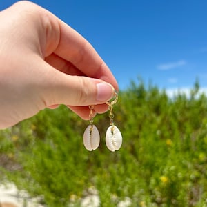 Peut inclure: Une paire de boucles d'oreilles en forme de coquillage dorées avec un charme en coquillage de cauris blanc. Les boucles d'oreilles pendent de la main d'une personne sur fond de feuillage vert et de ciel bleu.