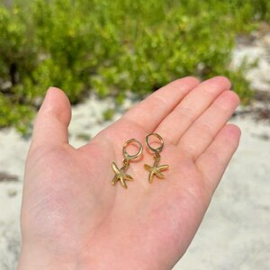 Könnte beinhalten: Ein Paar goldfarbene Seestern-Ohrringe. Jeder Ohrring hat einen kleinen, detaillierten Seestern-Anhänger, der an einem Reifen hängt. Die Ohrringe werden in einer Hand gehalten, mit einem Strand und Grün in der Hintergrund.