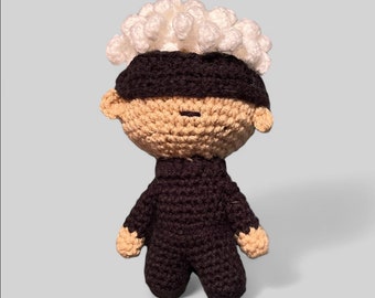 May include: A crocheted toy figure with a white crocheted hair, a black crocheted eye mask, and a brown crocheted body.