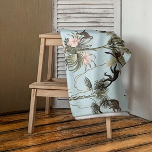 May include: A light blue blanket featuring a tropical design with monkeys, flowers, and palm leaves. The blanket is draped over a wooden stool, showcasing the detailed illustration and soft texture. The design includes pink flowers and brown monkeys.