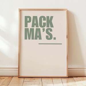 Könnte beinhalten: Gerahmter Kunstdruck mit dem Text "PACK MA'S." in Salbeigrün. Der Text ist auf einem cremefarbenen Hintergrund zentriert, mit einer horizontalen Linie darunter. Der Rahmen ist helles Holz, und der Druck wird auf einem Holzboden präsentiert.