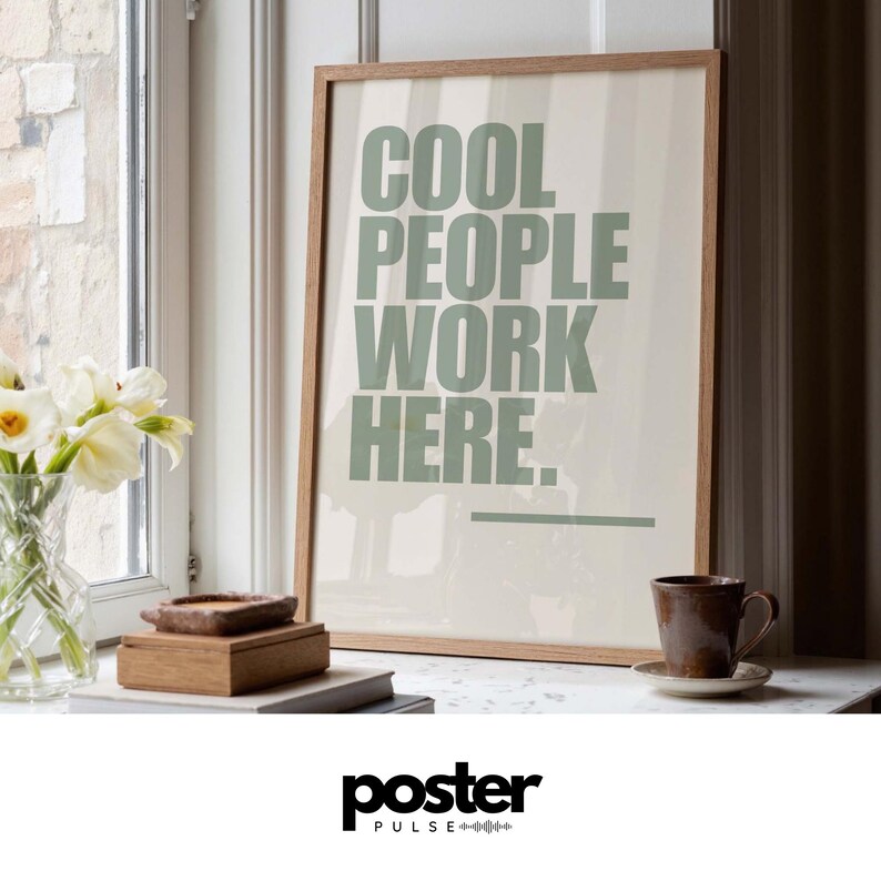 May include: A framed print with the text "COOL PEOPLE WORK HERE" in green letters. The print is in a light brown wooden frame. A brown ceramic mug sits on a white saucer next to the print.