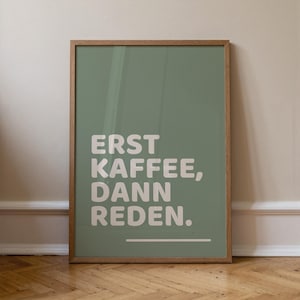Könnte beinhalten: Ein gerahmtes Wandbild mit einem salbeigrünen Hintergrund und dem weißen Text "ERST KAFFEE, DANN REDEN." sowie einer horizontalen weißen Linie darunter. Der Rahmen besteht aus hellbraunem Holz und hat ein schlichtes, rechteckiges Design.