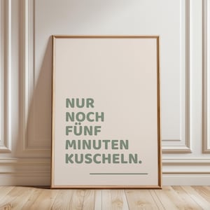 Könnte beinhalten: Ein rechteckiges Wandbild in einem hellen Holzrahmen mit cremefarbenem Hintergrund, das den salbeigrünen Text "NUR NOCH FÜNF MINUTEN KUSCHELN." sowie eine horizontale Linie unter dem Text in einer klaren, serifenlosen Schriftart zeigt.
