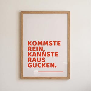 Könnte beinhalten: Ein gerahmter Druck mit dem deutschen Text "KOMMSTE REIN, KANNSTE RAUS GUCKEN" in roten Buchstaben auf weißem Hintergrund. Der Rahmen ist aus hellem Holz gefertigt. Unter dem Text befindet sich eine rote Linie.