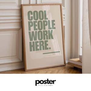 May include: A framed poster with the text "COOL PEOPLE WORK HERE" in green letters. The poster has a light beige background and a wooden frame. The poster is leaning against a white wall and a wooden floor.