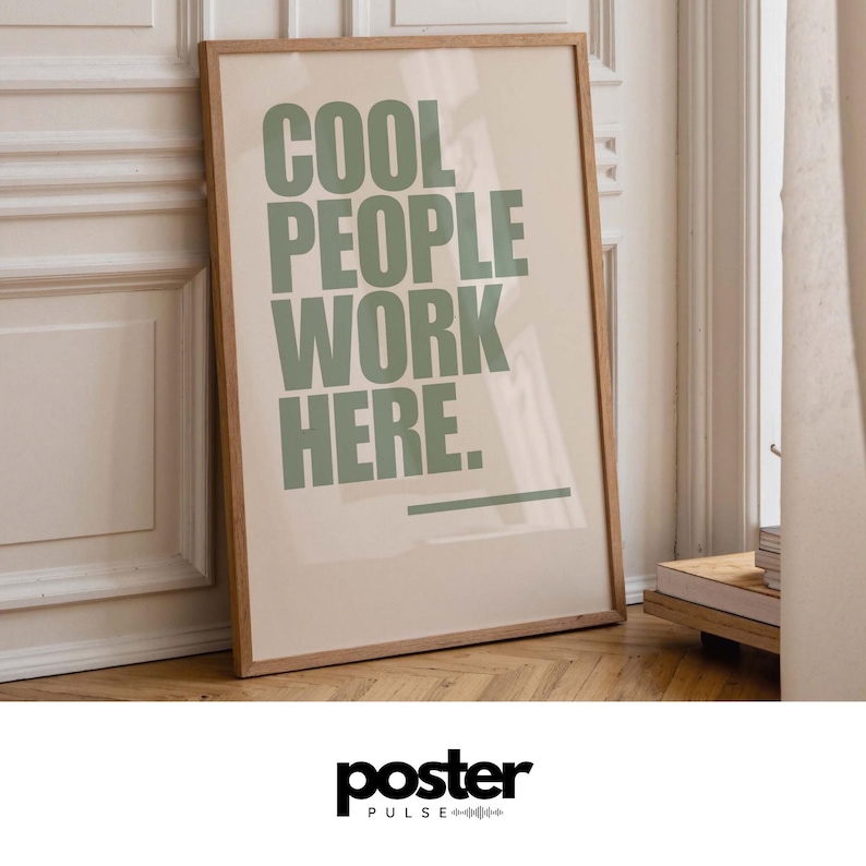 May include: A framed poster with the text "COOL PEOPLE WORK HERE" in green letters. The poster has a light beige background and a wooden frame. The poster is leaning against a white wall and a wooden floor.