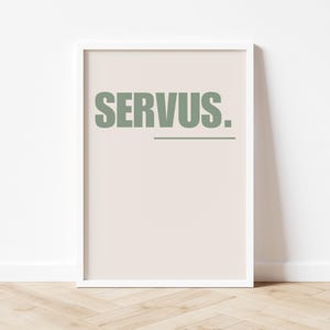 Op de afbeelding: Een witte geframed poster met een licht beige achtergrond en het woord "SERVUS." gedrukt in donkergroen.