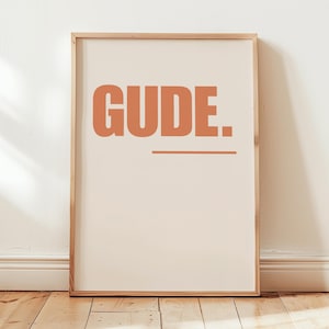 Peut inclure: Une affiche encadrée avec le mot "GUDE" en lettres orange. Le mot est au-dessus d'un trait horizontal, le tout sur un fond crème. Le cadre est en bois clair, et l'affiche est présentée sur un sol en bois.
