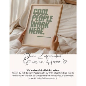 May include: A framed poster with the text "COOL PEOPLE WORK HERE" in green lettering. The poster is held by a person. The bottom of the poster has text in German.
