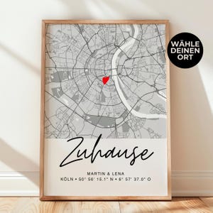 Puede incluir: Un mapa en blanco y negro de Colonia, Alemania, con un corazón rojo que marca el centro. El texto "Zuhause" está escrito en cursiva debajo del mapa, junto con los nombres "Martin & Lena" y las coordenadas "KÖLN 50° 56' 15.1 N 6° 57' 37.0 O". El texto "WÄHLE DEINEN ORT" está en un cuadro blanco con borde negro en la esquina superior derecha.
