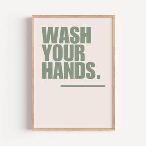 Op de afbeelding: Een ingelijste print met de woorden "WASH YOUR HANDS" in groene blokletters op een lichtroze achtergrond. Onder de tekst staat een dunne groene lijn. De lijst is lichtbruin.