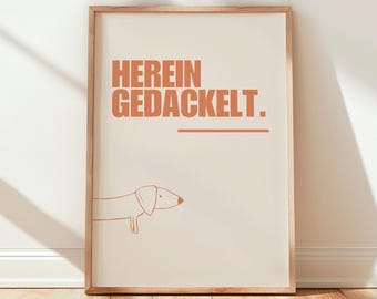 Può includere: Stampa artistica incorniciata con il testo tedesco "HEREIN GEDACKELT" in arancione, una linea e un'illustrazione minimalista di un bassotto. La stampa ha uno sfondo color crema e una cornice in legno.