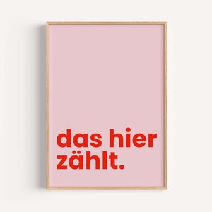 Könnte beinhalten: Gerahmter Druck mit hellrosa Hintergrund und dem deutschen Satz "das hier zählt" in roten Buchstaben. Der Holzrahmen verleiht dem minimalistischen Design eine natürliche Note. Geeignet für die Wohn- oder Bürodekoration.