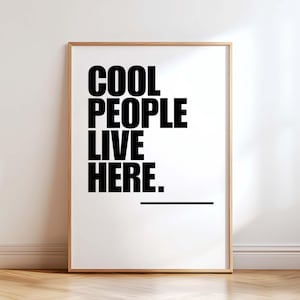 Può includere: Stampa incorniciata con la scritta "COOL PEOPLE LIVE HERE" in grassetto nero su sfondo bianco. Sotto il testo c'è una linea nera orizzontale. La cornice è di colore marrone chiaro.