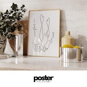 May include: A framed art print featuring a minimalist line drawing of three hands holding each other. The names "Paul", "Sara", and "Luna" are written in cursive. The artwork is in a gold frame and is displayed on a white surface.