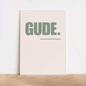 Könnte beinhalten: Ein weißer gerahmter Poster mit dem Wort "GUD.E." in hellgrün auf cremefarbenem Hintergrund.