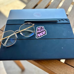 May include: A black Samsung tablet with a stylus and gold-rimmed glasses. A sticker with the text "NO PASA NADA" is on the tablet. A laptop is behind the tablet, resting on a wooden table.