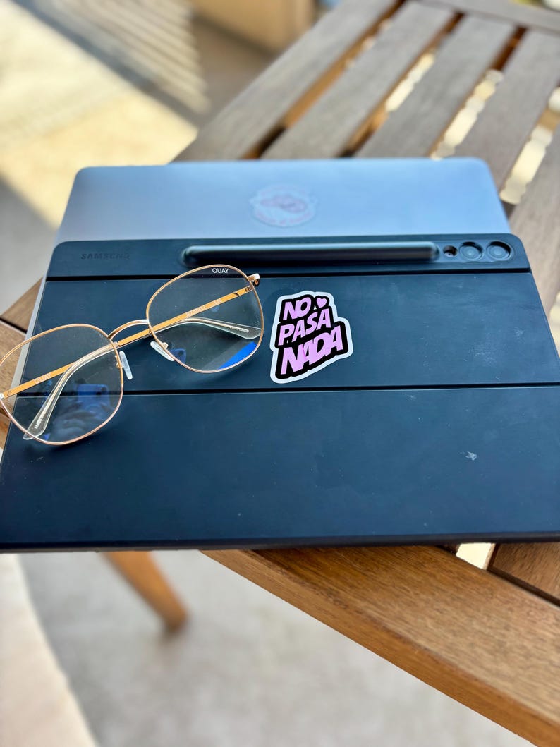 May include: A black Samsung tablet with a stylus and gold-rimmed glasses. A sticker with the text "NO PASA NADA" is on the tablet. A laptop is behind the tablet, resting on a wooden table.
