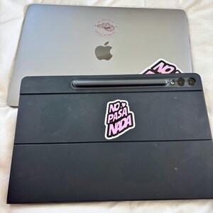May include: A silver laptop with a pink sticker that says "may had me at prince of paradise" and a black tablet with a pink sticker that says "NO PASA NADA". The tablet has a built-in stylus and camera.