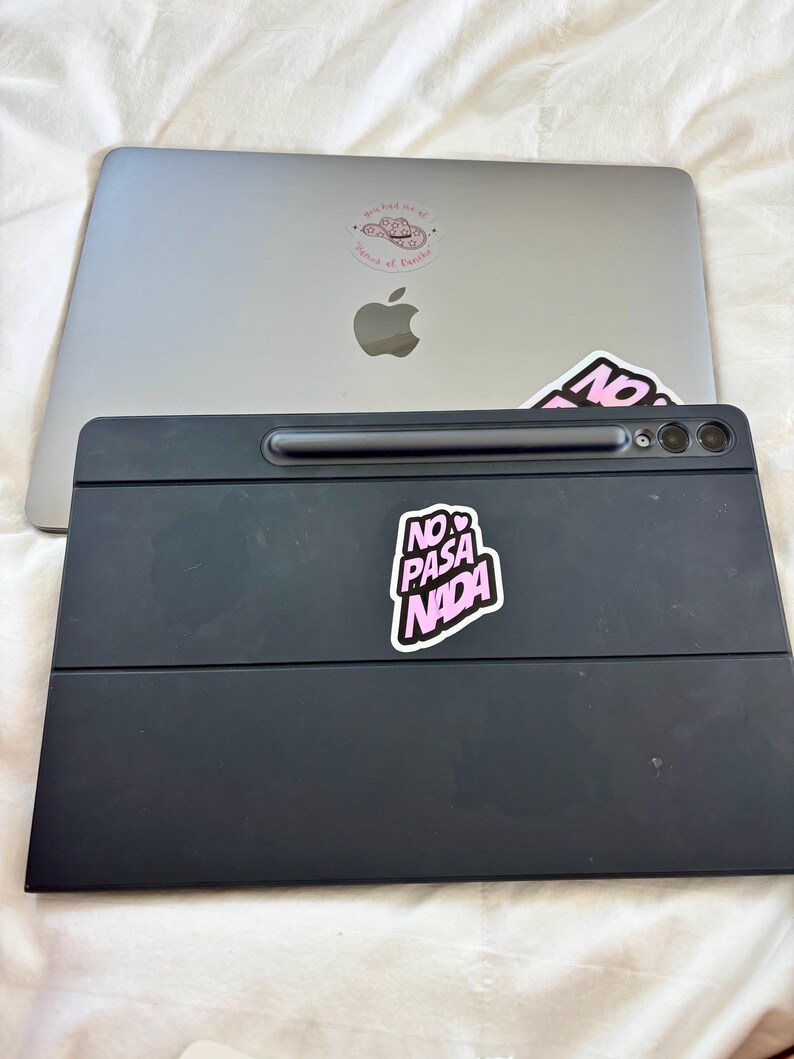 May include: A silver laptop with a pink sticker that says "may had me at prince of paradise" and a black tablet with a pink sticker that says "NO PASA NADA". The tablet has a built-in stylus and camera.