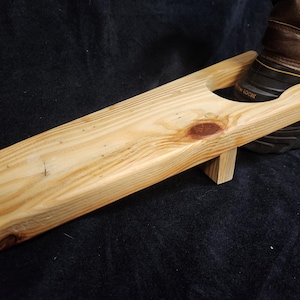 May include: A wooden boot jack made of light-colored pine. The boot jack has a long, flat body with a curved end and a circular opening. A brown boot is partially visible in the background.