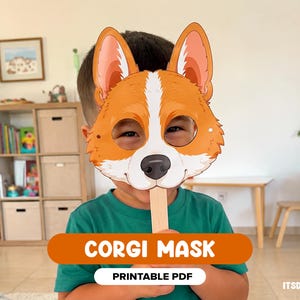 May include: A handheld Corgi mask featuring a brown and white dog face with large ears, a black nose, and eye cutouts, attached to a wooden stick. The mask includes the text CORGI MASK and PRINTABLE PDF on an orange and white banner overlay.