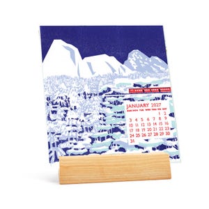 Puede incluir: Un calendario de escritorio para enero de 2027 con una ilustración estilizada de un tren rojo que viaja a través de un paisaje montañoso nevado con picos blancos y árboles azules, sostenido por un soporte de madera clara.