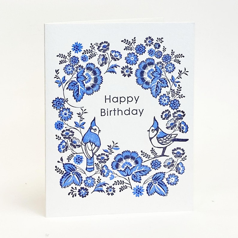 Może przedstawiać: Biała kartka urodzinowa z niebieskim wieńcem kwiatowym i dwoma niebieskimi ptakami. Słowa "Happy Birthday" znajdują się na środku. Kwiaty i ptaki są w odcieniach niebieskiego, tworząc dekoracyjny wz&oacute;r.