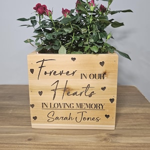 May include: A wooden planter box with the text "Forever in our Hearts" and "In loving memory Sarah Jones" engraved on it. The planter is filled with a small red rose bush.