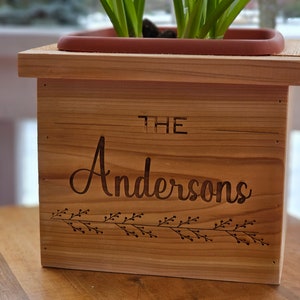 May include: A wooden planter box with the engraved text "THE Andersons" and a decorative floral design. The box is filled with soil and a plant. The planter is made of light-colored wood and sits on a wooden surface.
