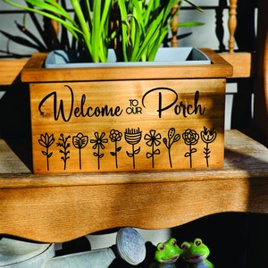 May include: Wooden planter box with the text "Welcome to our Porch" and a row of stylized flowers. The planter is sitting on a wooden bench.