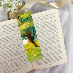 Puede incluir: Un marcapáginas caprichoso con una rana verde con un sombrero amarillo, sobre un fondo de un árbol y un campo de hierba. El marcapáginas está colocado en un libro abierto con texto.