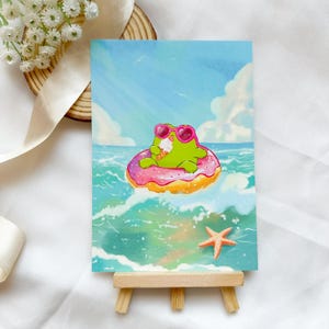 May include: A colourful illustration of a green frog wearing heart-shaped sunglasses, floating on a pink donut in the sea. The frog is holding ice cream. A starfish is in the lower right corner. The background is a blue sky and ocean.