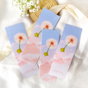 Puede incluir: Cuatro marcapáginas coloridos con un diseño de diente de león y rana. Los marcapáginas tienen un fondo de cielo rosa y azul con nubes blancas. El texto en los marcapáginas dice "Un libro es un billete a cualquier lugar que sueñes".