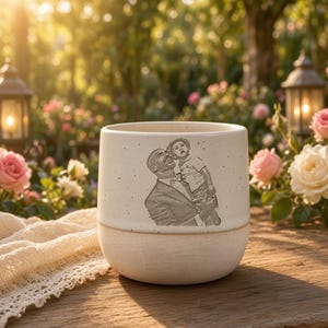 Könnte beinhalten: Ein Keramikbecher mit zweifarbigem Design, bestehend aus einem glatten, cremeweißen oberen Teil und einem strukturierten, matten beigen Boden. Auf der Vorderseite befindet sich eine detaillierte, dunkelgraue Gravur eines Erwachsenen mit Kind.