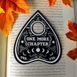 Może przedstawiać: Czarna i biała plansza Ouija z napisem "ONE MORE CHAPTER" i "YES" na dole. Plansza znajduje się na otwartej książce z jesiennymi liśćmi.