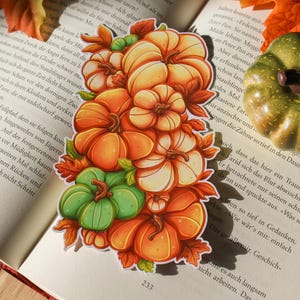 Könnte beinhalten: Ein Aufkleber mit einer Ansammlung von Kürbissen in Orange, Creme und Grün, mit Herbstblättern. Der Aufkleber befindet sich auf einem offenen Buch mit Text. In der Nähe liegt ein kleiner, grüner Kürbis.
