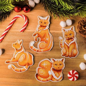 Könnte beinhalten: Eine festliche Anordnung mit orangefarbenen Katzenaufklebern mit weißen Akzenten, einer Zuckerstange und Weihnachtsschmuck. Die Aufkleber zeigen Katzen in verschiedenen Posen, mit einem Tannenzapfen und Grün.