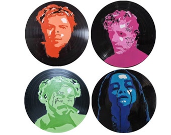 May include: Four vinyl records with painted portraits. The portraits are in orange, pink, green, and blue. The subjects are depicted in a stylized, artistic manner. The records are black with visible grooves.